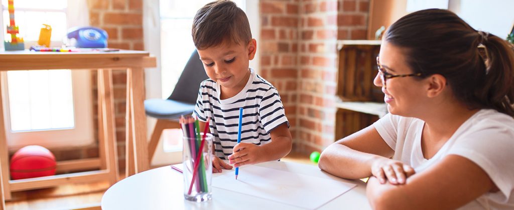 Young boy drawing with a colored pencil while a teacher looks on.